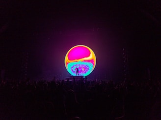 A vibrant, colorful circular screen dominates the center of the image, displaying swirling psychedelic patterns in hues of pink, yellow, and blue. In the foreground, the silhouette of a crowd is visible, suggesting the setting of a concert or live event.