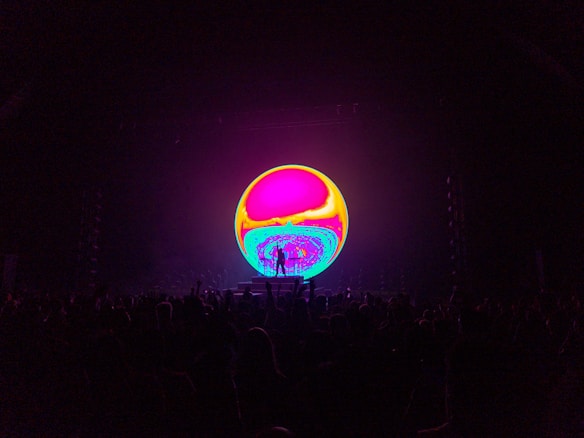 A vibrant, colorful circular screen dominates the center of the image, displaying swirling psychedelic patterns in hues of pink, yellow, and blue. In the foreground, the silhouette of a crowd is visible, suggesting the setting of a concert or live event.