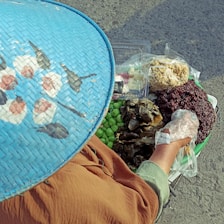 A person is wearing a blue woven hat with floral patterns and is handling food items in a basket. The basket contains various foodstuffs, including green fruit, wrapped items, and what appears to be different grains or rice. The person is using a plastic glove and is on a textured grey surface.