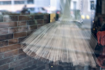 A delicate white ballet tutu with a fitted bodice embellished with light pearls and lace details. The tutu is displayed behind a glass window, showing reflections of the surroundings. A background of brown brick wall adds contrast to the elegant costume.