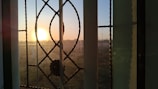 A decorative window with intricate patterns frames a bright, glowing sunset. The sun is low on the horizon, casting a warm light and creating shadows on the window. The glass has a floral embellishment and is divided into sections by metal lines.