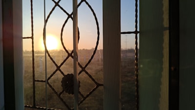 A decorative window with intricate patterns frames a bright, glowing sunset. The sun is low on the horizon, casting a warm light and creating shadows on the window. The glass has a floral embellishment and is divided into sections by metal lines.