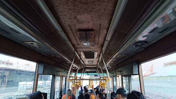 The interior of a bus is visible, showing the ceiling with patterned design and handrails. Several passengers are seated or standing, and there are windows providing a view of the airport tarmac and a parked airplane. The no smoking signs are prominently displayed.