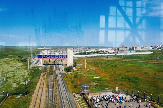 A vast landscape with railway tracks running towards a large sign that reads 'РОССИЯ', indicating the border with Russia. The foreground shows green fields and a crowd of people standing on a platform. Buildings and infrastructure are visible in the background under a clear blue sky.