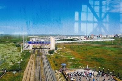A vast landscape with railway tracks running towards a large sign that reads 'РОССИЯ', indicating the border with Russia. The foreground shows green fields and a crowd of people standing on a platform. Buildings and infrastructure are visible in the background under a clear blue sky.