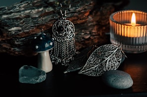 An arrangement of items featuring a small ceramic mushroom with a dark blue cap speckled with white, a piece of translucent crystal, and intricately designed metallic jewelry shaped like leaves and a dreamcatcher. These objects are placed on a surface with a textured wooden log in the background, and a lit candle in a glass holder casting a warm glow over the scene.
