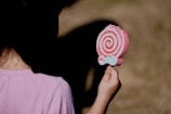 A child wearing a pink shirt is holding a lollipop-shaped ice cream with pink and white swirls under bright sunlight. The background is blurred with earthy tones, and the focus is on the colorful treat.