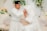 A bride and groom are seated on an elegant couch, adorned with light-colored pillows. The groom, dressed in white traditional attire with a black hat, is gently kissing the bride's hand. The bride is wearing a white dress with lace details and a long veil, and her hands are decorated with intricate henna designs. Behind them, there are floral arrangements featuring green leaves and white and peach flowers.