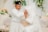A bride and groom are seated on an elegant couch, adorned with light-colored pillows. The groom, dressed in white traditional attire with a black hat, is gently kissing the bride's hand. The bride is wearing a white dress with lace details and a long veil, and her hands are decorated with intricate henna designs. Behind them, there are floral arrangements featuring green leaves and white and peach flowers.