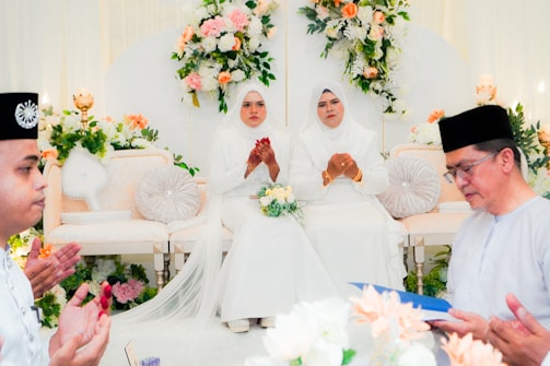 Four people are gathered in what appears to be a ceremonial setting. Two women in white attire are seated on an ornate sofa, surrounded by elaborate floral decorations. They have their hands clasped in front, suggesting they might be praying or participating in a religious or cultural ritual. Two men are sitting or standing nearby, one reading from a blue book or document.