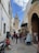 A narrow, cobblestone street flanked by historic buildings, bustling with people and small shops. A tall minaret stands prominently in the distance, indicating an Islamic architectural style. The sky is partly cloudy, and various items are on display along the street, including crafts and food items.