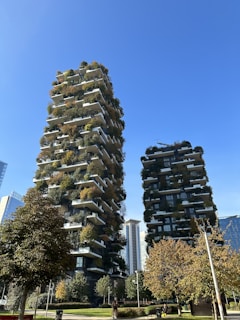 Two tall buildings are covered with lush greenery, incorporating plants and trees on multiple balcony levels. A clear blue sky serves as the backdrop while trees and a person walking in a park are visible in the foreground.