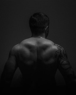 A black and white photograph of a muscular man shown from the back. He has short hair and a prominent tattoo on his upper right arm, extending partially down the arm. The lighting emphasizes the well-defined muscles of his back and shoulders.