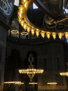 Large, ornate chandeliers hang from a high ceiling in an opulent interior, illuminated with warm light. Arabic calligraphy adorns the walls in a richly detailed and historical architectural setting.