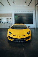 A vibrant yellow sports car is showcased in a modern, minimalistic showroom. The car is facing forward, displaying its sleek and aerodynamic front design with sharp headlights. The background features a large digital screen and a display cabinet with various items.