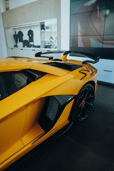 A sleek yellow sports car with a prominent rear wing is parked indoors. The image captures part of the car's rear and side, highlighting its aerodynamic features and glossy finish. Behind the car, a wall-mounted showcase displays various merchandise, including black shirts and accessories. The setting appears to be a modern showroom with clean lines and a minimalist design.