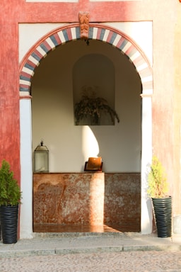 A decorative architectural archway with alternating black and white stripes at the top. Below the arch, there is an alcove with a brown marble base. Inside the alcove, a book is displayed on a wooden stand, illuminated by sunlight. On either side of the alcove, two potted plants in black planters are present.