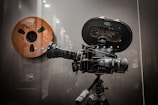A vintage film reel and an old-fashioned Arriflex film camera mounted on a tripod are displayed against a reflective surface. The camera is equipped with a large lens and various knobs, showcasing intricate mechanical details.