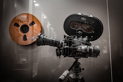 A vintage film reel and an old-fashioned Arriflex film camera mounted on a tripod are displayed against a reflective surface. The camera is equipped with a large lens and various knobs, showcasing intricate mechanical details.