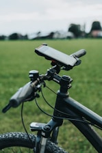 A close-up view of a bicycle handlebar equipped with a smartphone mount and a small device attached to the frame. The bicycle is positioned on a grassy field, with a blurred background of trees and a cloudy sky.