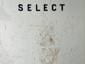 Black capital letters spelling 'SELECT' are mounted on a textured wall with a rough, abstract pattern. The wall features various shades of off-white with hints of beige and brown markings scattered randomly.