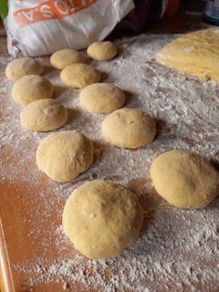 Round balls of dough are scattered across a floured surface. A bag of flour is partially visible in the background, and a larger portion of dough sits nearby.