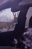 A small, clear glass bottle with a wooden cap is hanging from the rearview mirror of a vehicle. The vehicle window appears to be fogged or frosted, and the environment outside is somewhat obscured, suggesting cold weather.