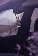 A small, clear glass bottle with a wooden cap is hanging from the rearview mirror of a vehicle. The vehicle window appears to be fogged or frosted, and the environment outside is somewhat obscured, suggesting cold weather.