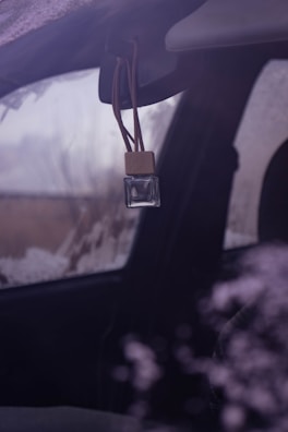 A small, clear glass bottle with a wooden cap is hanging from the rearview mirror of a vehicle. The vehicle window appears to be fogged or frosted, and the environment outside is somewhat obscured, suggesting cold weather.