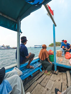 A small boat with several people sitting on wooden benches. One person is wearing a hat, and another is dressed in a brown headscarf and patterned pants. The boat is on calm water with more boats and a small island visible in the background. The Indonesian flag is attached to a post, and various items such as bags and a water jug are placed on the floor.