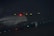 An airplane with the Star Alliance livery is positioned on a dimly lit runway at night. The red light near the aircraft's tail is illuminated, and several small green lights line the ground near the plane. The setting is dark with some faintly visible airport signage in the background.