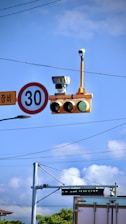 A traffic light with a green signal is mounted on a pole alongside a speed limit sign indicating 30. The setup is equipped with several surveillance cameras, situated against a backdrop of power lines and a clear blue sky with some clouds.