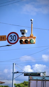 A traffic light with a green signal is mounted on a pole alongside a speed limit sign indicating 30. The setup is equipped with several surveillance cameras, situated against a backdrop of power lines and a clear blue sky with some clouds.
