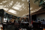 A cozy outdoor cafe setting with large, cream-colored canopy shades creating a dappled effect from sunlight filtering through nearby trees. Several people stand at a wooden counter, suggesting a busy, lively environment. Wooden furniture and green plants contribute to a natural, relaxed atmosphere.