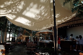 A cozy outdoor cafe setting with large, cream-colored canopy shades creating a dappled effect from sunlight filtering through nearby trees. Several people stand at a wooden counter, suggesting a busy, lively environment. Wooden furniture and green plants contribute to a natural, relaxed atmosphere.