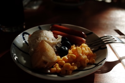 A plate of food featuring a variety of items, including corn, blueberries, a cooked sausage, and a piece of steamed fish. The plate is decorated with simple blue patterns and rests on a wooden table. The lighting creates shadows, providing a warm and homey atmosphere.