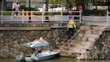 A person wearing a life jacket is climbing a stone staircase by a waterway. There is a small boat tied to the steps, with orange cones visible on the sidewalk above. Several people are walking on the sidewalk, and there are trees and foliage in the background.