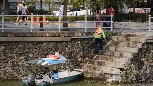 A person wearing a life jacket is climbing a stone staircase by a waterway. There is a small boat tied to the steps, with orange cones visible on the sidewalk above. Several people are walking on the sidewalk, and there are trees and foliage in the background.