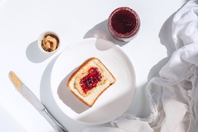 A slice of bread with peanut butter and jelly spread on a white plate. Nearby, there is an open jar containing red jelly and a small bowl with peanut butter. A butter knife with peanut butter rests on the side, and a crumpled white napkin is visible in the corner.