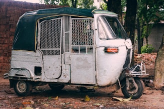 A rustic, weathered auto rickshaw with a metal cage covering stands parked on a dirt path. Surrounding the vehicle are trees and scattered leaves, and a brick wall can be seen in the background.