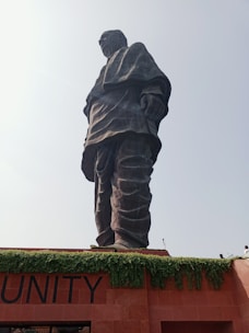 A towering statue stands prominently against a bright sky. The figure is draped in a long garment and appears to be made of dark stone or metal, conveying a sense of dignity and strength. Below the statue, greenery drapes over the edges of a reddish-brown structure, with the word 'UNITY' partially visible.