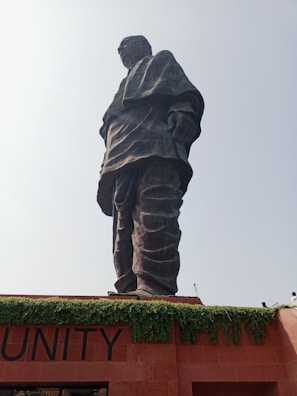 A towering statue stands prominently against a bright sky. The figure is draped in a long garment and appears to be made of dark stone or metal, conveying a sense of dignity and strength. Below the statue, greenery drapes over the edges of a reddish-brown structure, with the word 'UNITY' partially visible.