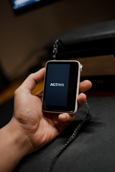 A hand is holding a small, portable electronic device with a screen displaying the word 'ACTIVO'. The device is connected to a black cable.