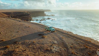 A 4x4 vehicle navigating rocky coastal terrain with waves crashing nearby under clear skies.