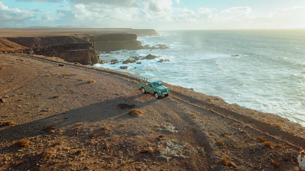A 4x4 vehicle navigating rocky coastal terrain with waves crashing nearby under clear skies.