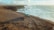 A rugged off-road vehicle is parked on a windswept, rocky coastline under partly cloudy skies. The ocean waves crash against the cliffside, creating a dramatic scene with a sense of adventure and isolation.