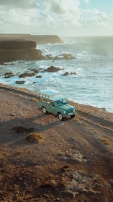 A vintage off-road vehicle is parked on a rocky coastal landscape beside the ocean, with waves crashing against the cliffs and a partly cloudy sky above.