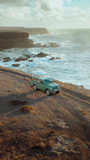 A vintage off-road vehicle is parked on a rocky coastal landscape beside the ocean, with waves crashing against the cliffs and a partly cloudy sky above.