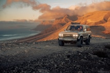 A four-wheel-drive vehicle is driving on a rocky, gravel road in a mountainous area during sunset. The background features rugged mountains, a coastline, and a cloudy sky with warm hues of orange and yellow. The scene exudes a sense of adventure and exploration.