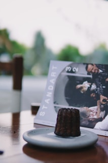A close-up view of a dark, textured pastry on a light gray plate set on a wooden table. In the background, there is a magazine titled 'Standart' featuring a group of people engaged in a conversation. The setting appears to be outdoors with blurred greenery in the distance.